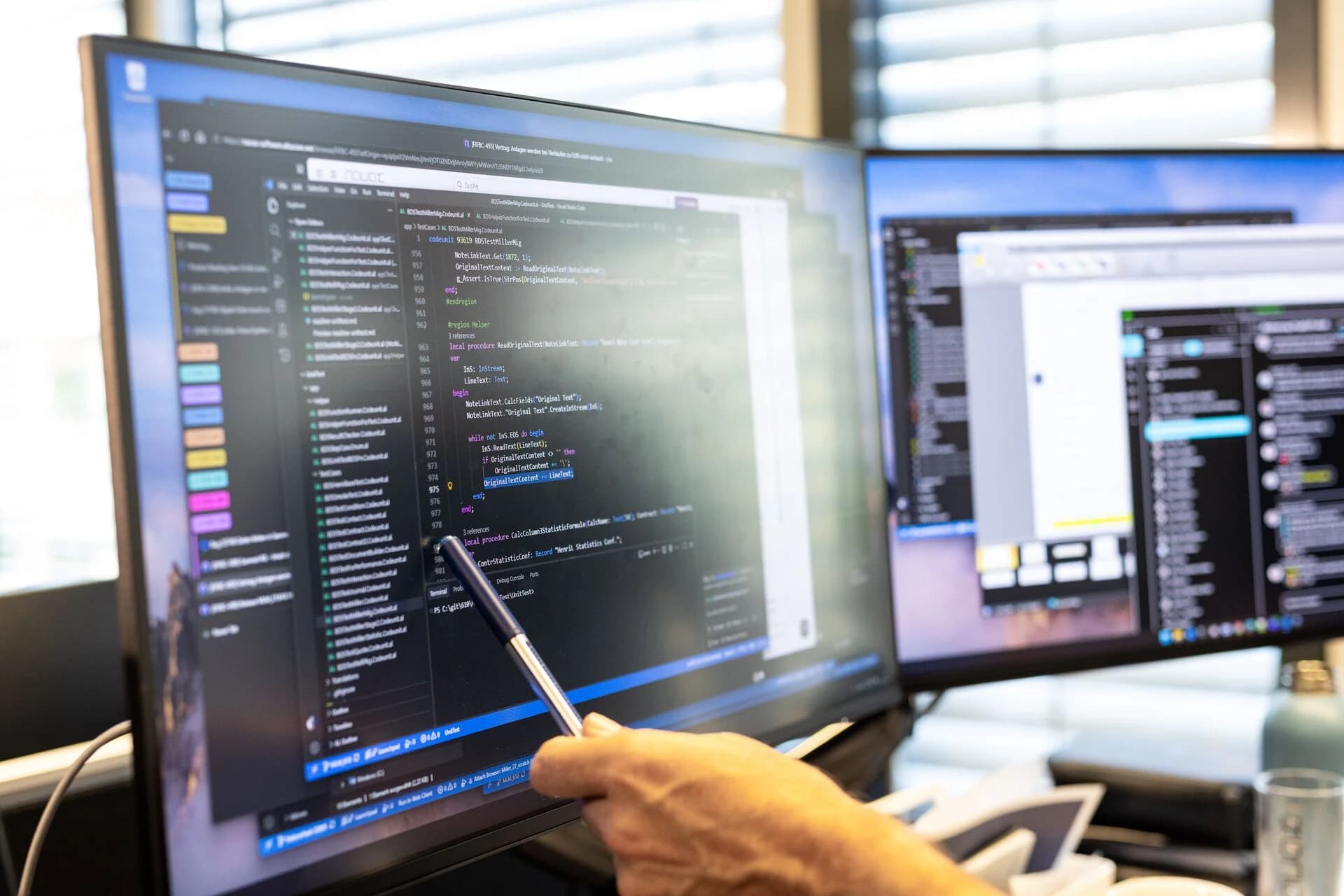Bildschirme mit Programmcode, eine Hand zeigt mit einem Stift darauf