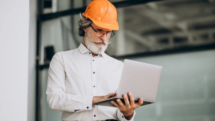 Mann trägt Bauhelm und hält Laptop in der Hand 