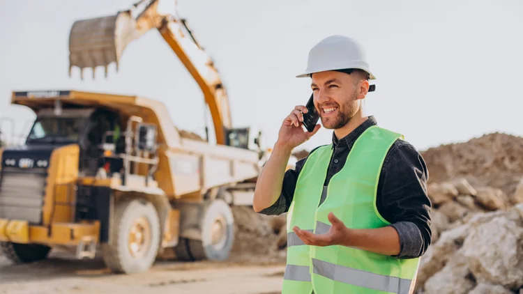 Mann steht vor Maschinen auf Baustelle und telefoniert 